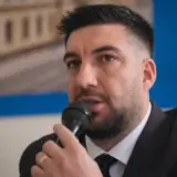 A man in a suit speaking into a microphone with text on the background wall
