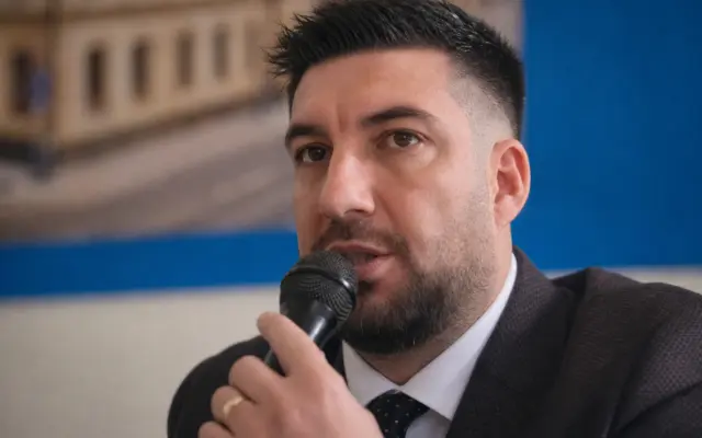A man in a suit speaking into a microphone with text on the background wall