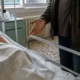 O persoană aflată pe un pat de spital, acoperită cu un cearșaf alb, este vizibilă din profil (partea brațului)