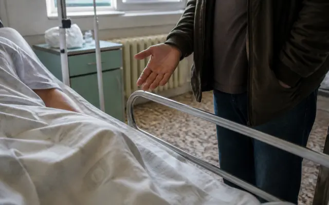 O persoană aflată pe un pat de spital, acoperită cu un cearșaf alb, este vizibilă din profil (partea brațului)