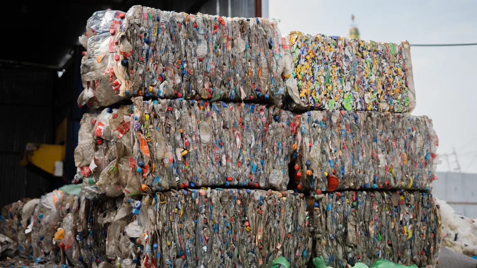 O stivă de baloturi compacte de deșeuri reciclabile, în special sticle de plastic și ambalaje de diverse culori, depozitate într-o hală de sortare