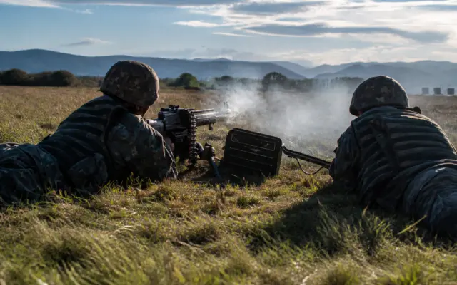 Two soldiers in camouflage uniforms operating a machine gun during military training in a grassy field