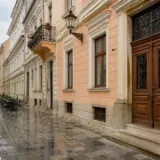 O stradă pietonală istorică, pavată cu piatră cubică umedă, mărginită de clădiri vechi cu arhitectură clasică în nuanțe de pastel