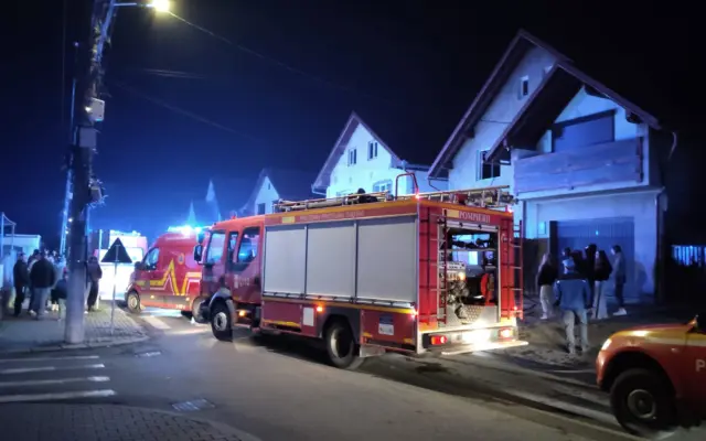 O fotografie de noapte care prezintă o autospecială de pompieri (ISU) și o ambulanță SMURD parcate pe o stradă rezidențială în fața unor case