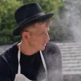 A man in traditional attire and apron stirring a large steaming cauldron outdoors at a museum