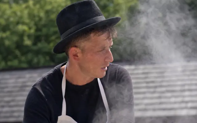 A man in traditional attire and apron stirring a large steaming cauldron outdoors at a museum