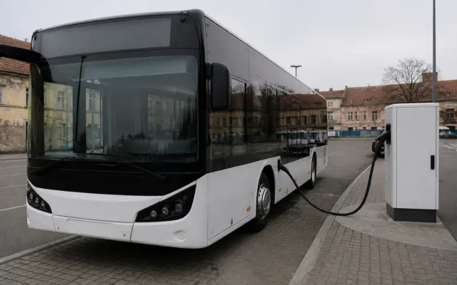 Un autobuz modern, alb cu negru, este parcat langa o statie de incarcare electrica alba intr-o zona urbana