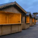 Mai multe căsuțe din lemn pentru târg, aliniate într-o piață publică, luminate cald la interior și decorate cu ghirlande verzi la streșini